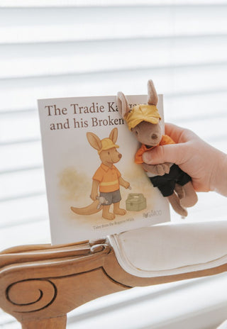 Hand holding a small plush toy next to a book titled 'The Tradie Kangaroo and his Broken Heart'.