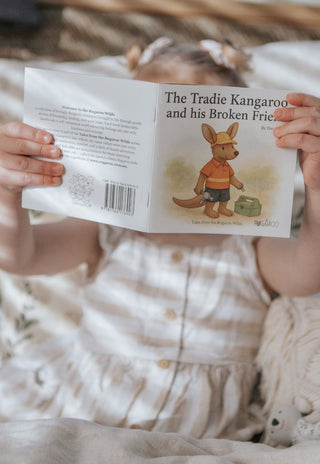 Child holding a book titled 'The Tradie Kangaroo and his Broken Frie' with a kangaroo illustration on a soft surface.