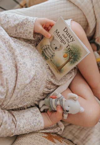 Child holding a book titled 'Dusty and the Mango Tree' and a plush koala toy, sitting on a couch.