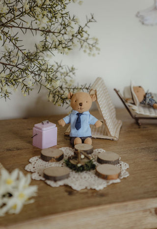 A small Rugaroo wombat Prep Kindy doll wearing a light blue shirt and dark tie sits on a wooden table, surrounded by miniature props including a pink esky, in a campground miniature dollhouse scene.