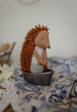A small echidna Rugaroo doll with brown yarn-like spines and a white striped shirt, sitting in a miniature tinnie boat made from metal.