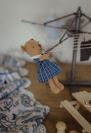 A small plush Rugaroo wombat prep/kindy doll in a school uniform dress hangs from a miniature clothesline in a styled room scene with a wooden floor, patterned fabric, and toy furniture.