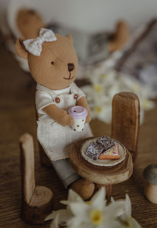 A miniature Rugaroo Barista Wombat doll wearing a white apron and bow, sitting at a miniature wooden table with a tiny cup and a slice of lamingtons and Fairy Bread, in a softly blurred rustic setting.