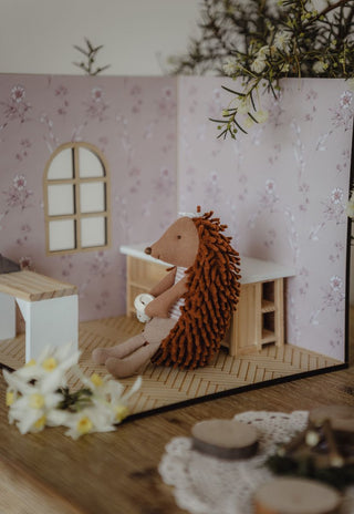 A small plush Rugaroo echidna doll with brown shaggy spikes and a beige body sits inside a miniature wooden playhouse with pink floral wallpaper, a window, and small furniture pieces.