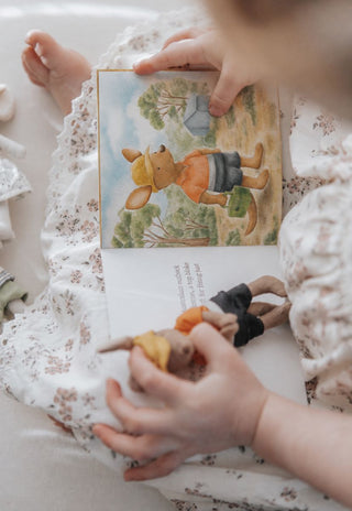 A close-up lifestyle shot of a child's hands holding an open children's book with a kangaroo illustration, surrounded by soft white floral fabric. The book is The Tradie Kangaroo and his Broken Friend by Rugaroo.