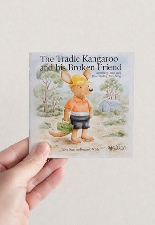 A hand holds a children's book titled "The Tradie Kangaroo and his Broken Friend" against a plain light beige background. The cover shows an illustrated kangaroo Tradie Rugaroo doll wearing a yellow hard hat, orange shirt, and dark shorts, standing outdoors near a small cabin.