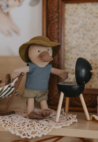 Small platypus plush doll wearing a hat and shirt, standing next to a miniature barbecue grill.