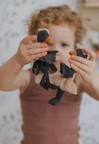 Young child holding up two black swan Rugaroo heirloom dolls; one in a black tulle skirt, one in a blush tutu, against a botanical wallpaper background