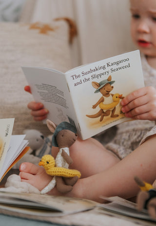 Child reading a Rugaroo children's book with a kangaroo illustration, surrounded by toys on a soft surface.