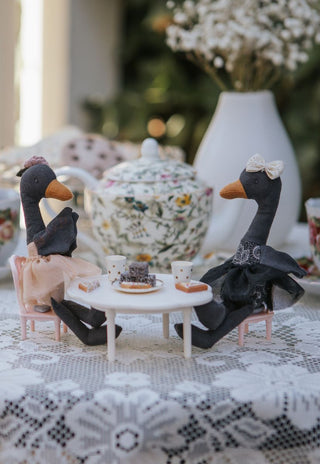 Two black swan Rugaroo dolls seated at a miniature tea party table with tiny cups, a lamington, and a floral teapot in the background