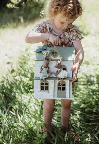 Child standing in long grass holding a sage green dollhouse with Mon the Bae and Betty the Bush Belle Rugaroo kangaroo dolls on the balcony