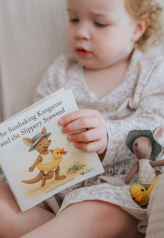Child reading a Rugaroo children's book with a kangaroo illustration, surrounded by toys on a soft surface.