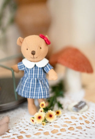 A Rugaroo miniature wombat kindy/prep doll in a blue school uniform, standing on a white lace doily with small yellow flowers and a mushroom-shaped object in a soft-focus background.