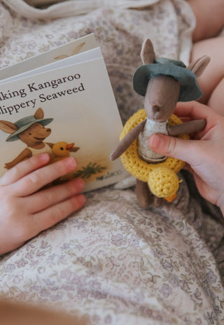 Child reading a Rugaroo children's book with a kangaroo illustration, surrounded by toys on a soft surface.