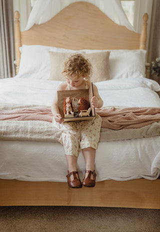 A young child with curly hair sits on a bed holding a small wooden dollhouse filled with miniature rugaroo echidna toys. 