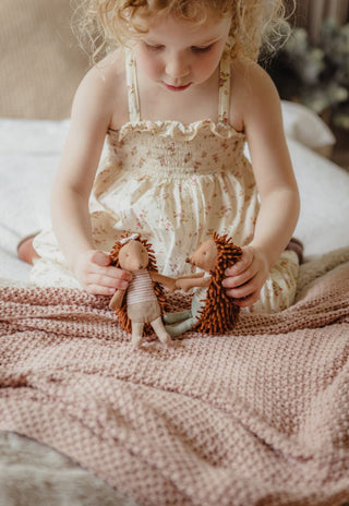 A young child sitting on a bed wrapped in a dusty pink knit blanket, holding and playing with Rugaroo's dollhouse scale Australian echidna dolls.
