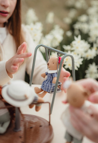 A close-up lifestyle shot of a girl interacting with a miniature Rugaroo wombat doll on a small swing, with a blurred background of white flowers.