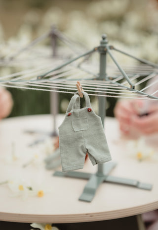 A miniature sage fabric clothing dryer hanging from a small clothesline stand on a table, with a tiny wooden clothespin holding the garment. The background is softly blurred with warm, neutral tones and hints of hands or objects out of focus.