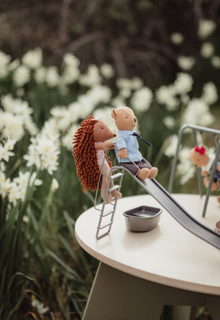 A styled product shot of a miniature doll playset on an outdoor patio. The set includes a dollhouse slide, ladder, small table, and plush toys, with a Rugaroo echidna on the ladder and a wombat sitting on the slide.