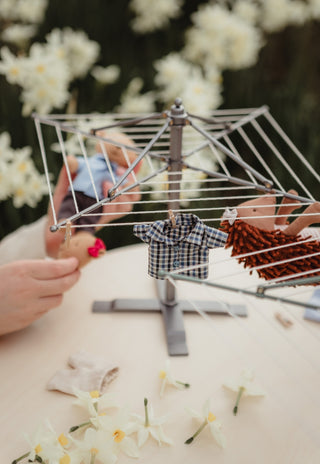 A miniature Rugaroo metal clothesline set up on a table with small doll-sized clothing items hanging on it, surrounded by blurred white flowers and hands adjusting the setup.