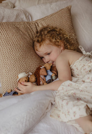 A young child with curly red hair lies on a bed with a textured beige pillow, holding rugaroo echidna and wombat miniature dolls.