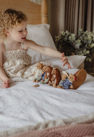 A young child in a cream floral nightgown sits on a bed with white bedding, reaching toward a group of miniature Rugaroo Australian echidna and wombat dolls.