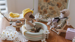 Two toy dollhouse platypus in a setting with a small pool and a chair, on a wooden surface.