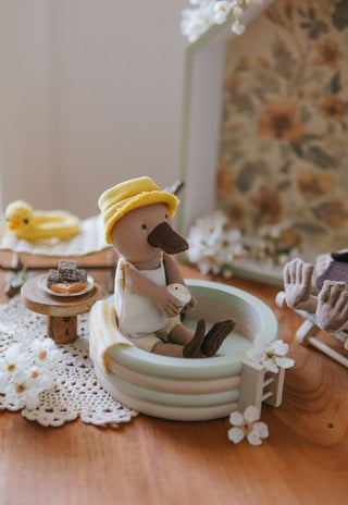Toy dollhouse platypus in a setting with a small pool and a chair, on a wooden surface.