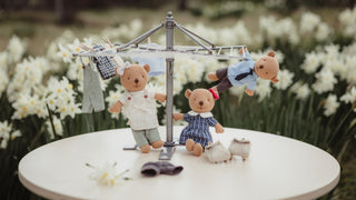 Three Australian Rugaroo wombat dolls dressed in miniature clothing are posed around a small sollhouse scene clothesline set on a round white table, with blurred white flowers in the background.