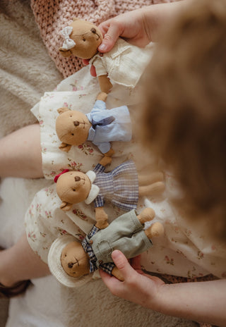 A close-up, top-down view of a child's hands holding four small Rugaroo wombat dolls dressed in different outfits, resting on a floral cream dress on a soft beige blanket.